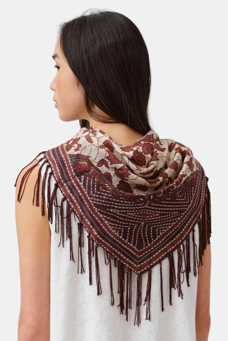 A woman shown from the back wearing a large fringed shawl scarf with a brown and beige floral pattern, paired with a white sleeveless top against a light gray studio background.