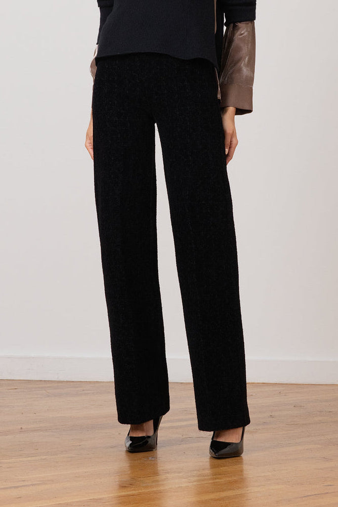 A woman stands in a studio wearing a black long-sleeve knit top with brown leather sleeves and black wide-leg trousers, paired with black pointed shoes.