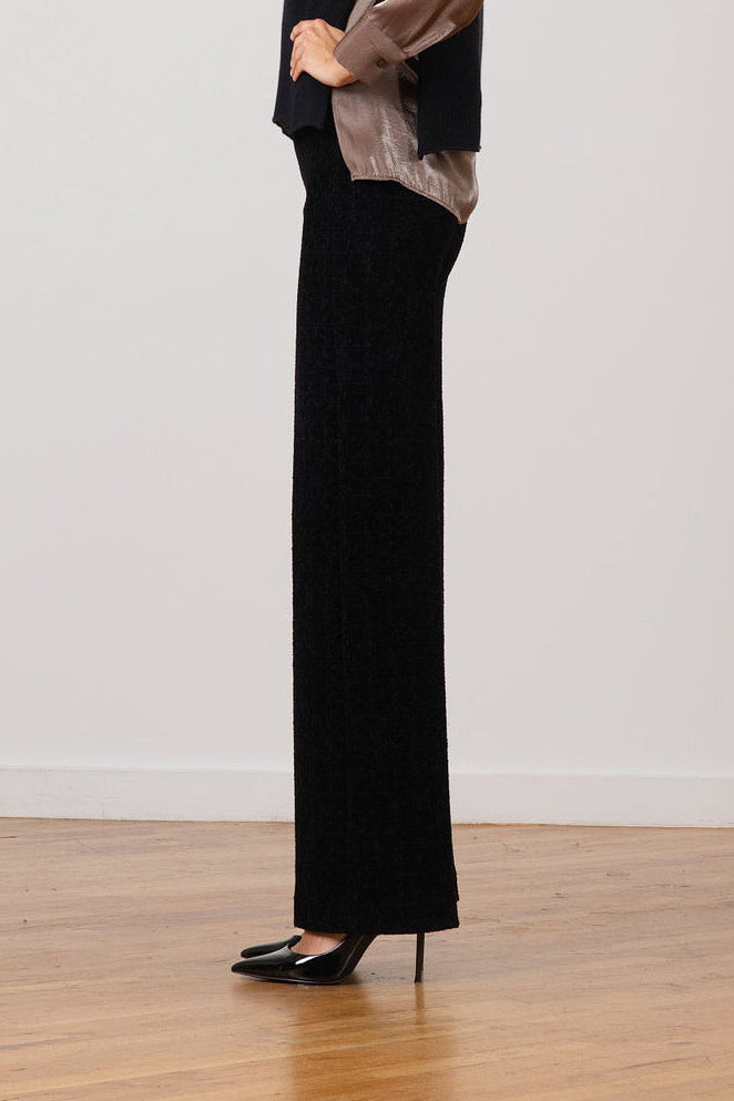 A woman stands in profile wearing a black long-sleeve top with sheer taupe sleeves and black wide-leg trousers, standing on a light wood floor against a plain off-white wall.