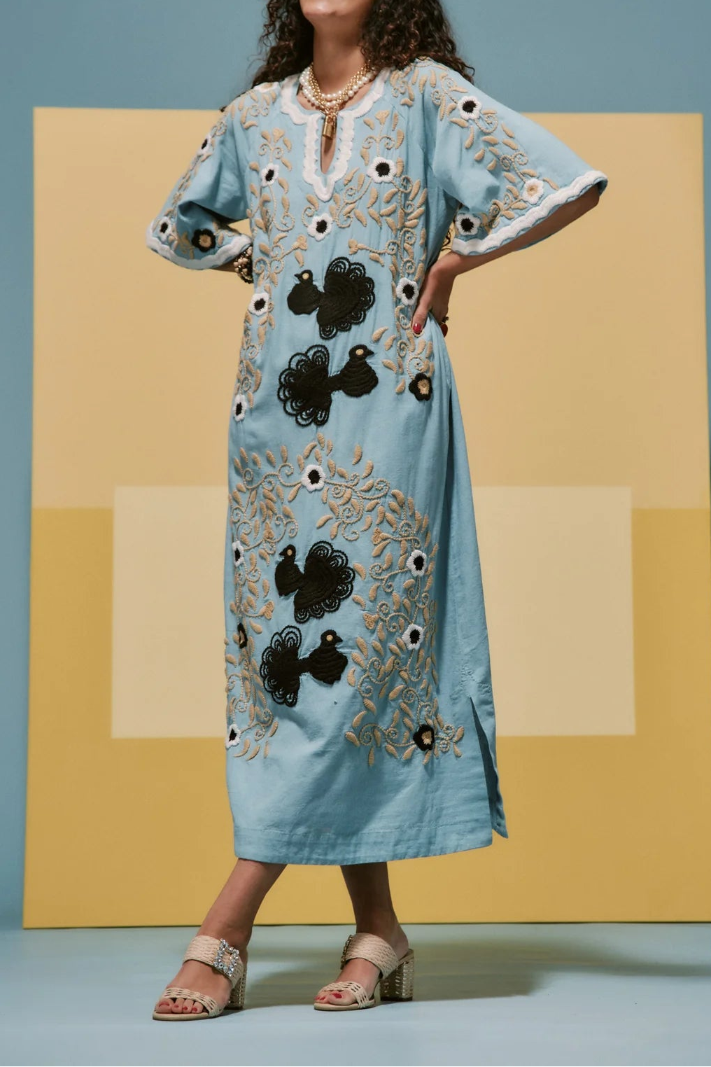 A woman modeling a light blue kaftan dress with black and gold floral embroidery, standing in front of a yellow geometric backdrop against a blue wall.