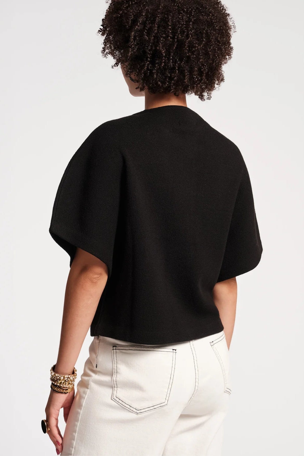 A person shown from the back wearing a black short-sleeve knit top and off-white pants against a plain light gray studio background.