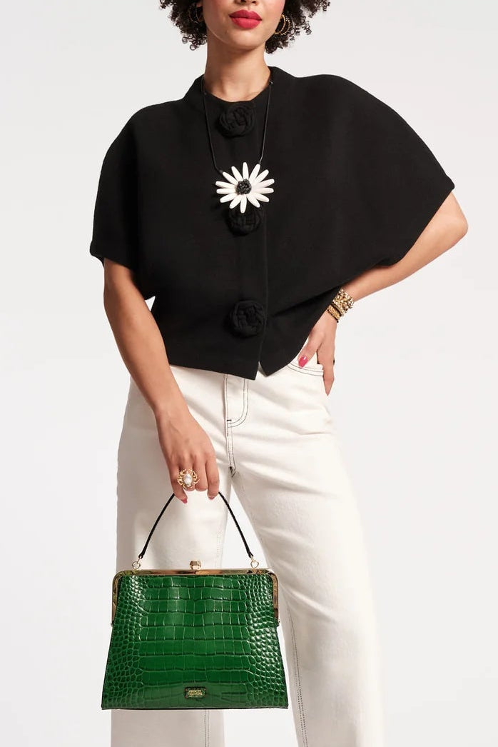 A model stands against a light gray studio background wearing a black short-sleeve blouse with a white starburst chest emblem, white wide-leg pants, and holding a green crocodile-embossed handbag.