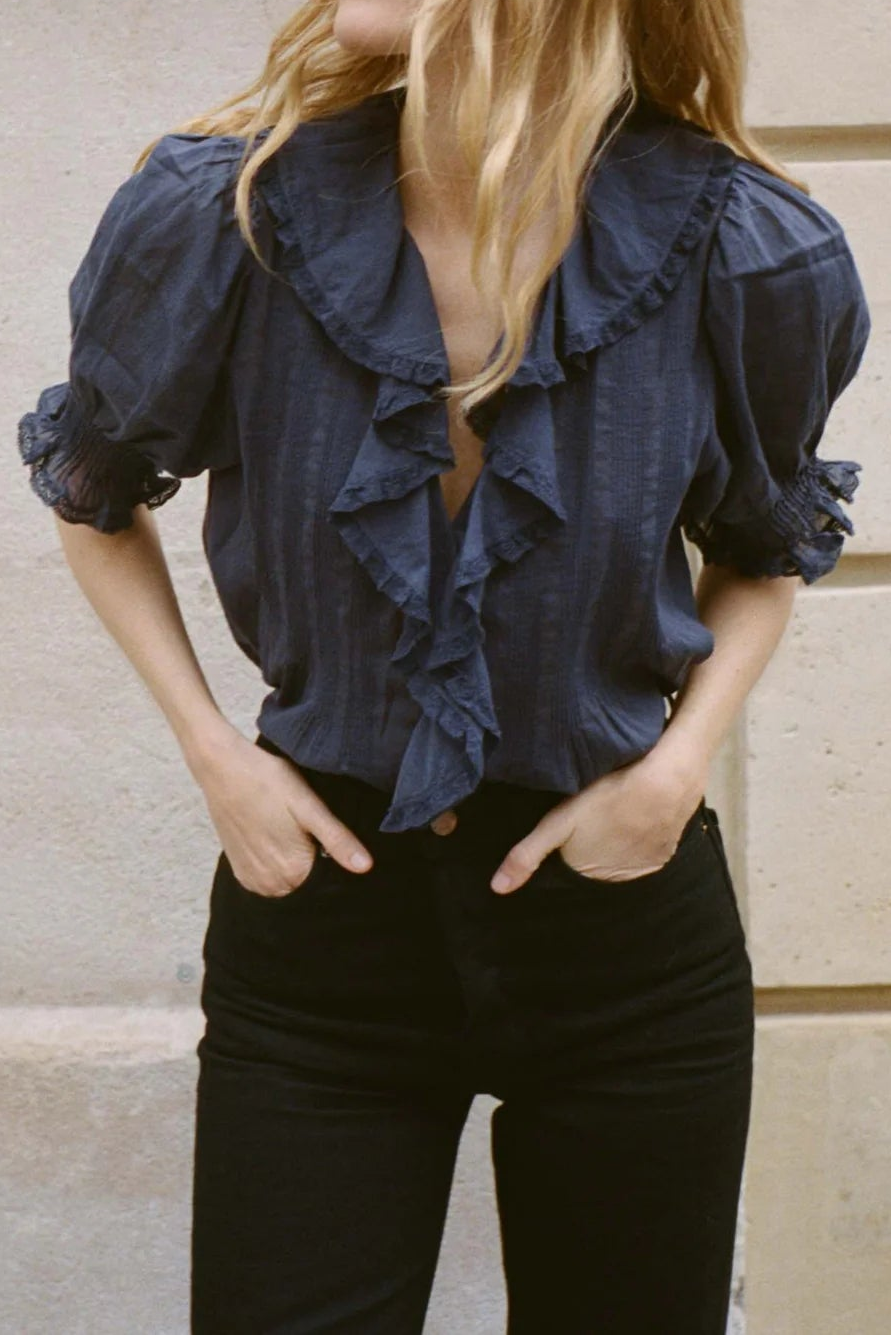 Woman wearing a navy blue blouse with ruffles against a beige wall.
