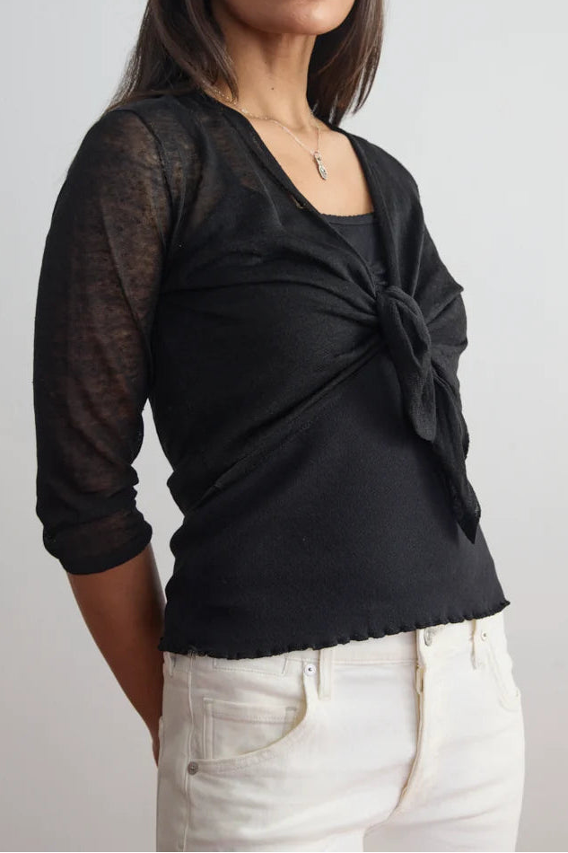 A woman modeling a black sheer three-quarter sleeve top with a twisted front detail, paired with white pants, against a plain light gray background.