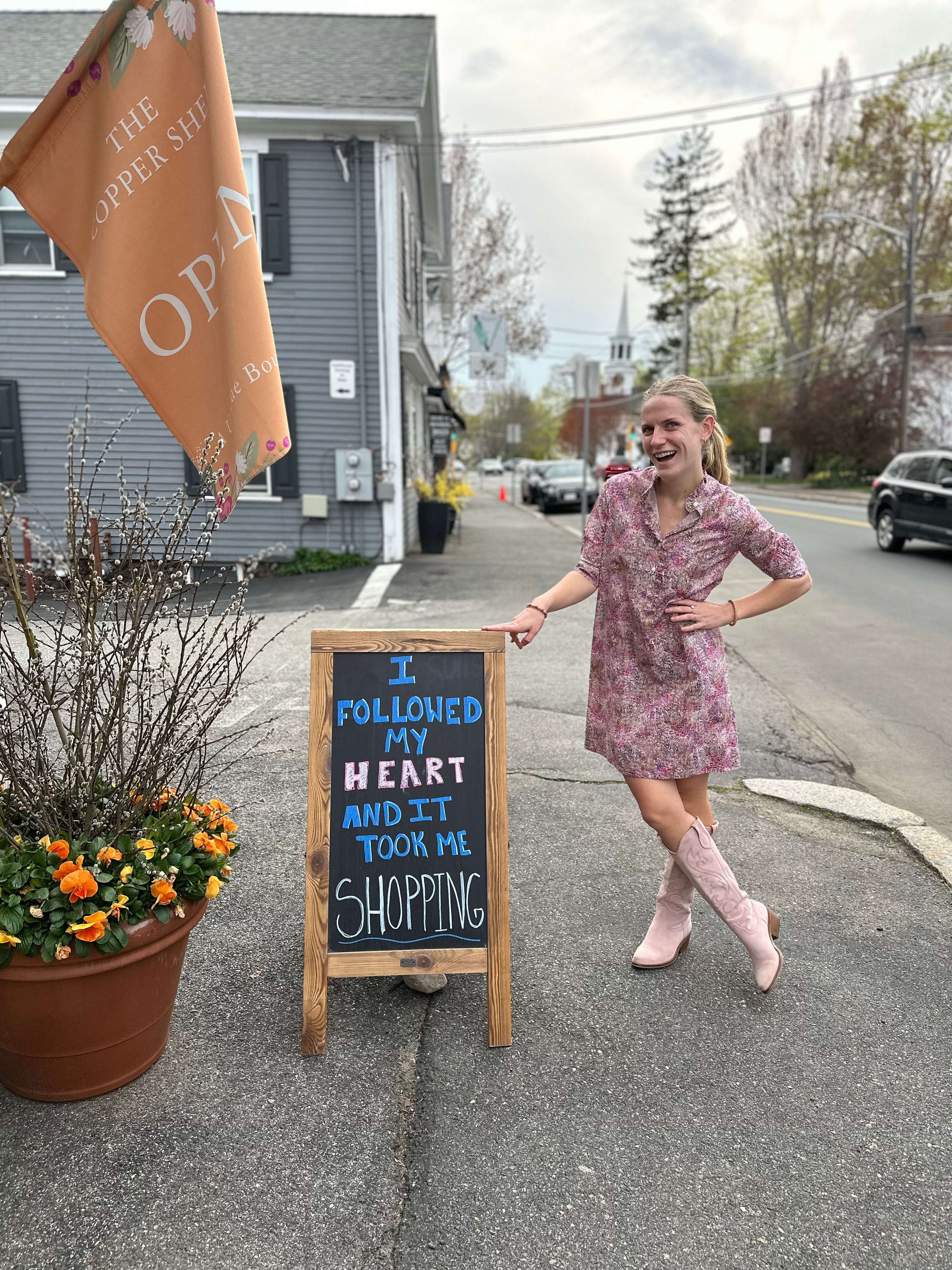 Woman standing next to a chalkboard sign on a street corner with a store sign in the background.