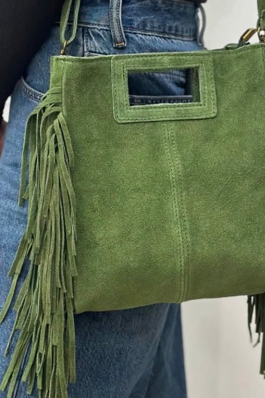 An olive green suede shoulder bag with long fringe and a rectangular cutout detail on the front, shown being worn over the shoulder of a person wearing blue jeans.
