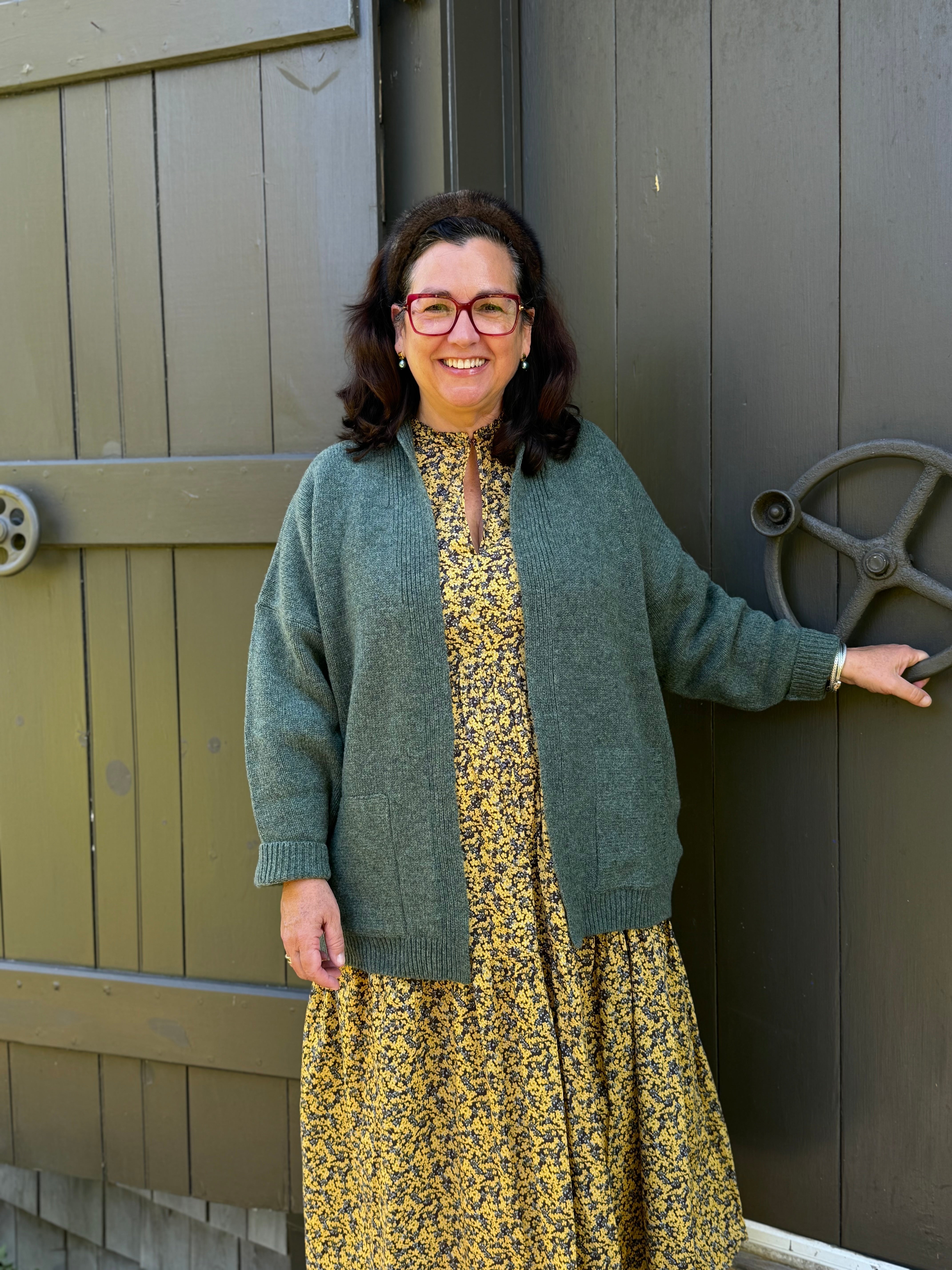 Woman wearing a green cardigan and yellow floral dress standing next to a wooden door.