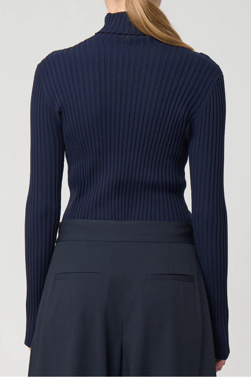 Person wearing a navy ribbed turtleneck sweater and dark pants on a light gray background