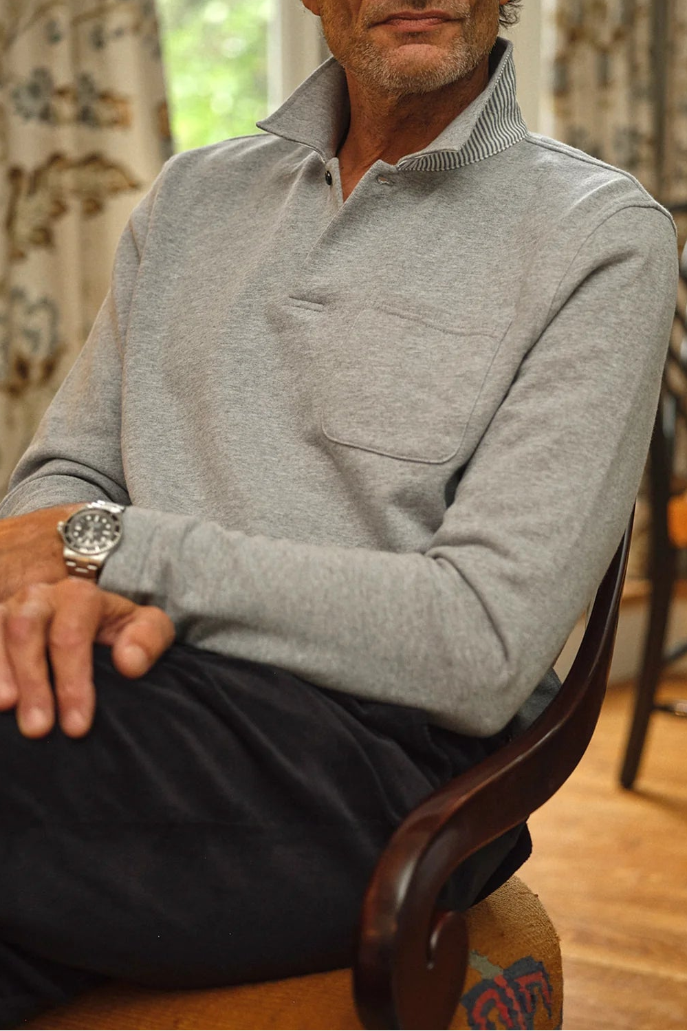 A man with graying hair and a beard sits in a wooden chair in a warmly lit room, wearing a light gray long-sleeve polo sweater and dark trousers. A patterned curtain, a window, and other furniture are visible in the background.