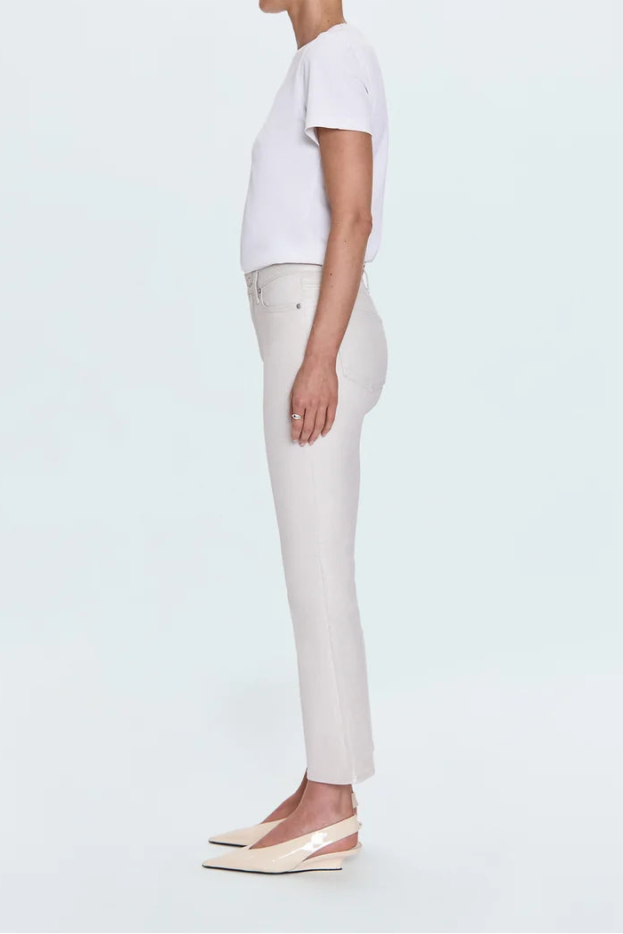 A woman in profile wearing a white short-sleeve top, light beige cropped trousers, and beige slingback wedge flats, standing against a very light gray studio background.