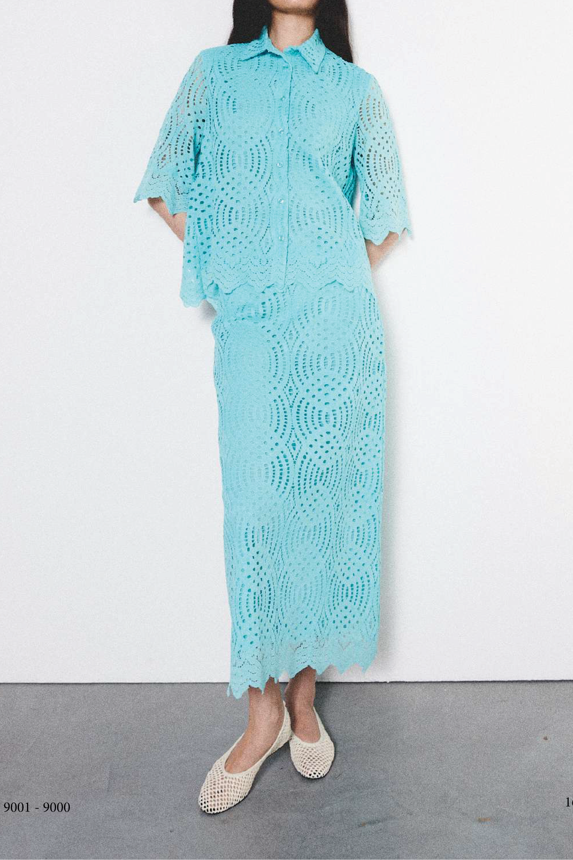 A model stands against a white wall wearing a light aqua blue crochet knit collared shirt and matching midi skirt set, paired with beige flat shoes.