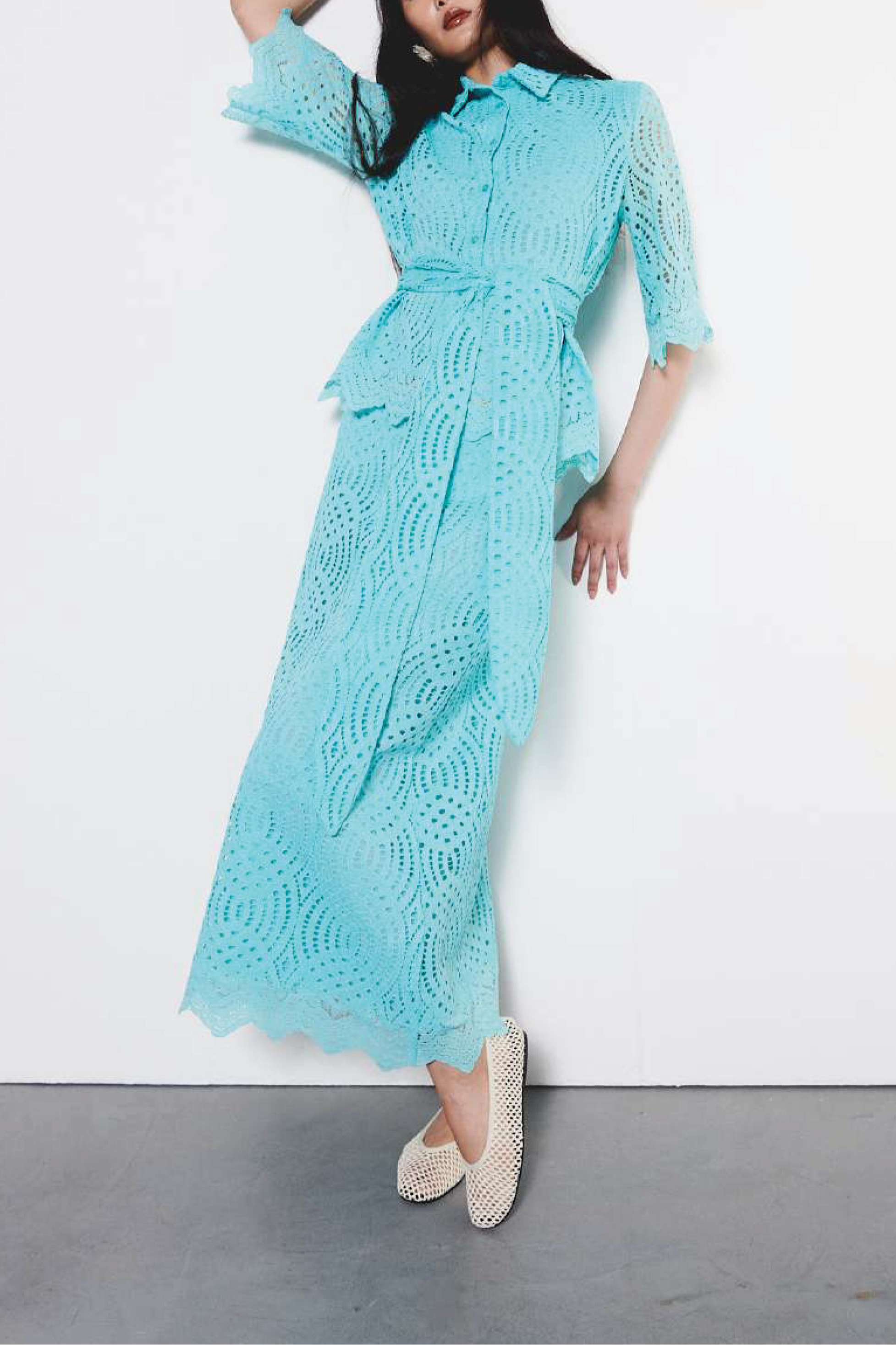A woman modeling a light blue crochet lace midi dress with a collar, button front, and matching tie belt, standing against a plain white wall with a gray floor.