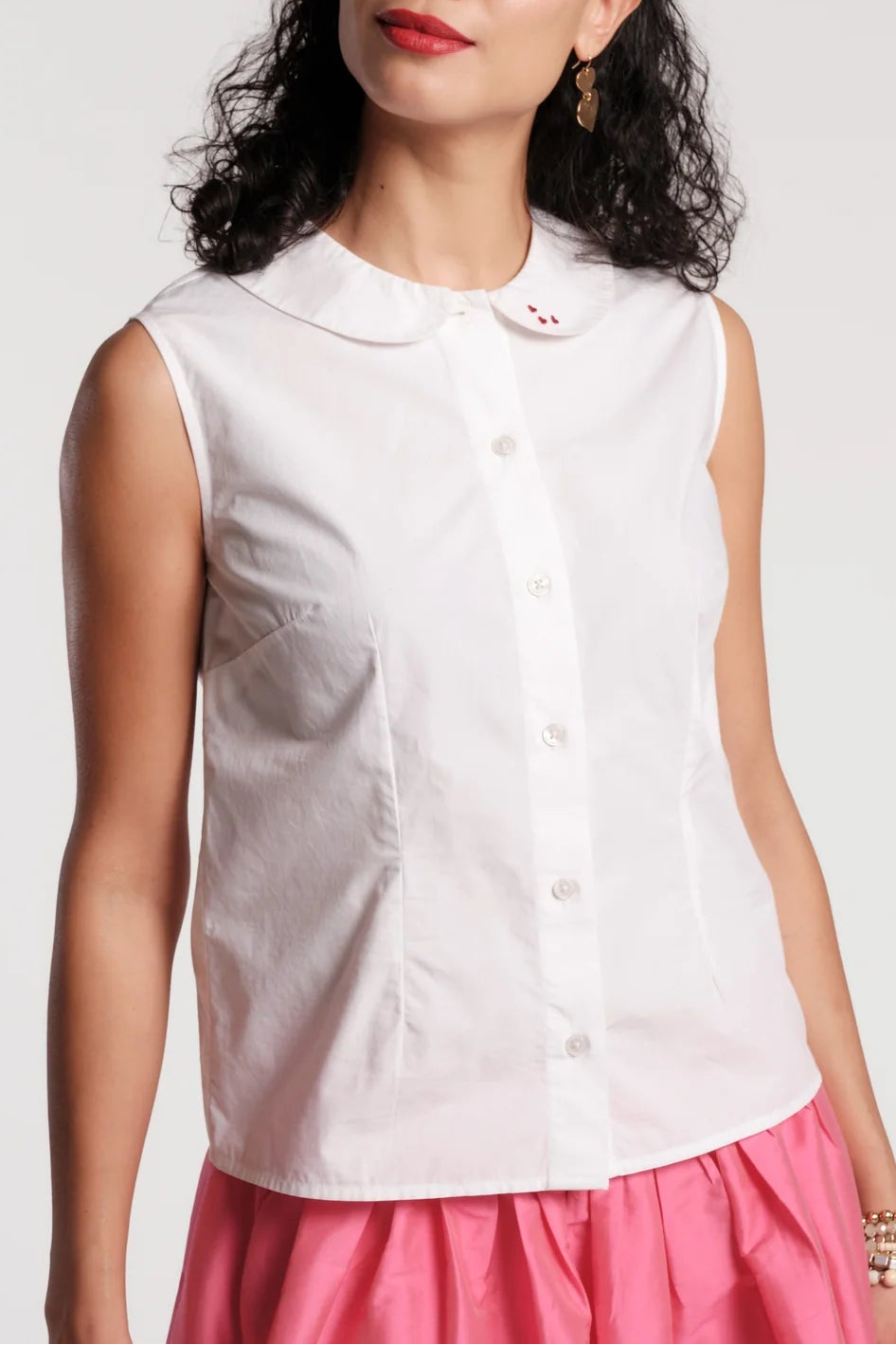 A woman wearing a sleeveless white button-front top with a Peter Pan collar and a pink skirt, photographed against a light gray studio background.