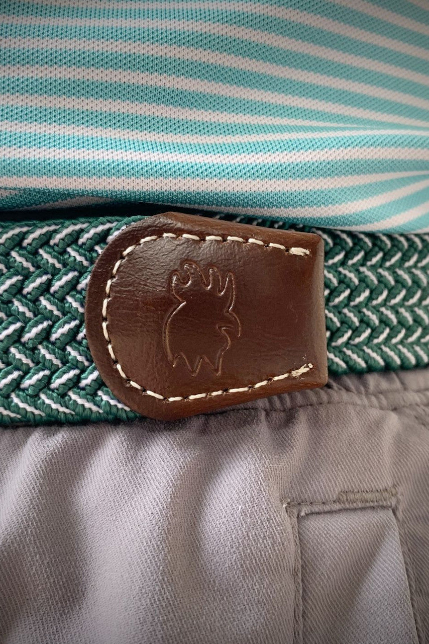 Close-up of a woven belt with a brown leather logo patch, worn around the waist over a striped shirt and gray pants.