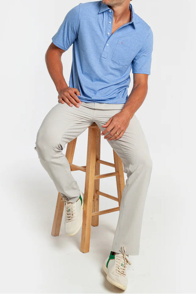 Man sitting on a wooden stool wearing a blue polo shirt and light gray pants against a white background