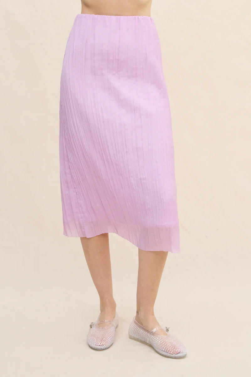 A model is wearing a light purple pleated midi skirt with a slight asymmetrical hem, paired with light-colored woven slip-on shoes, against a solid beige background.