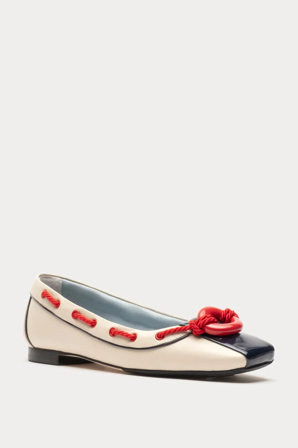 A single ballet flat shoe with a cream and navy color-blocked design, featuring red decorative stitching and a red knotted bow on the toe, set against a light gray background.