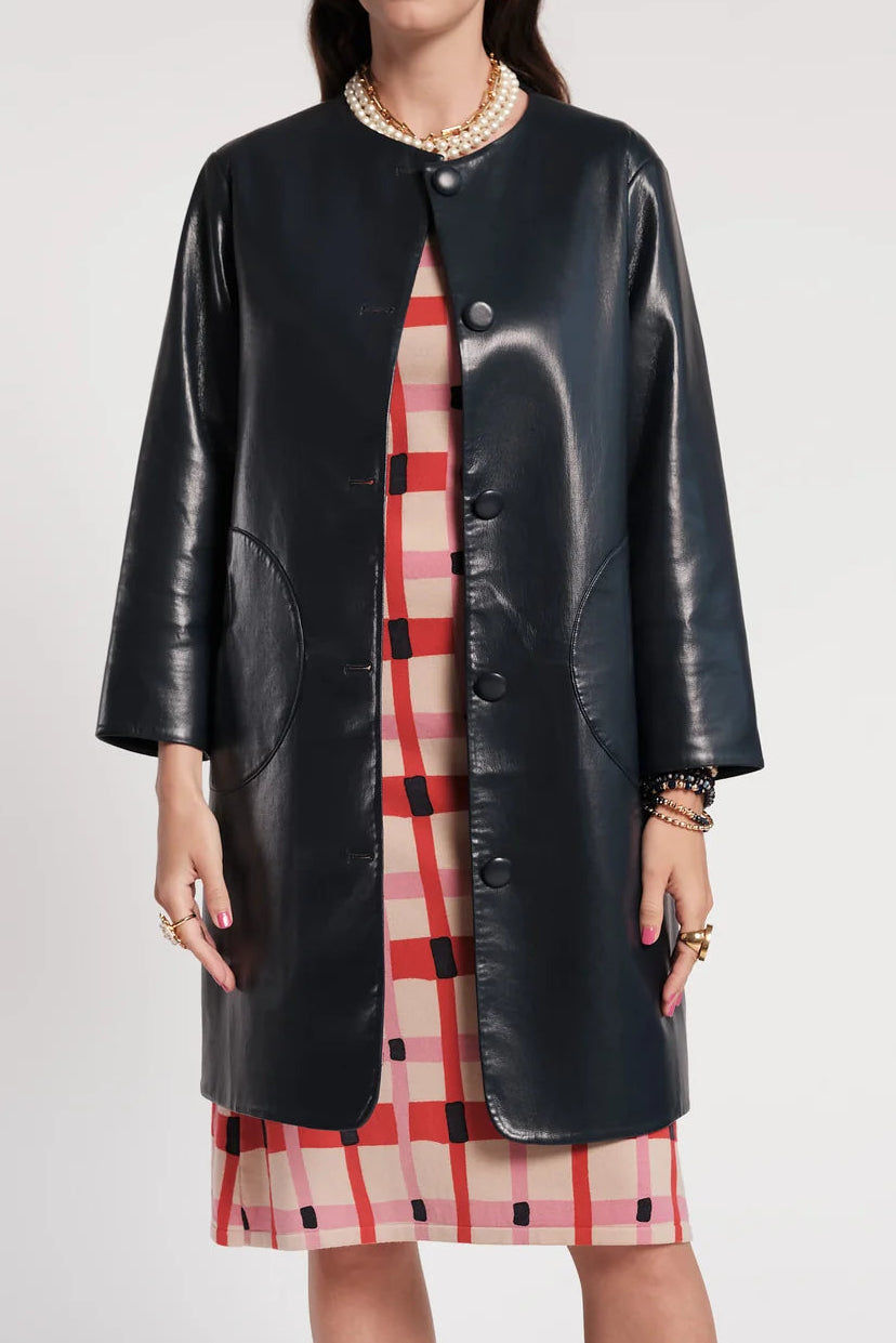 A model wearing a black faux-leather coat over a red, white, and black plaid dress, styled with a pearl necklace and gold jewelry, against a light gray studio background.