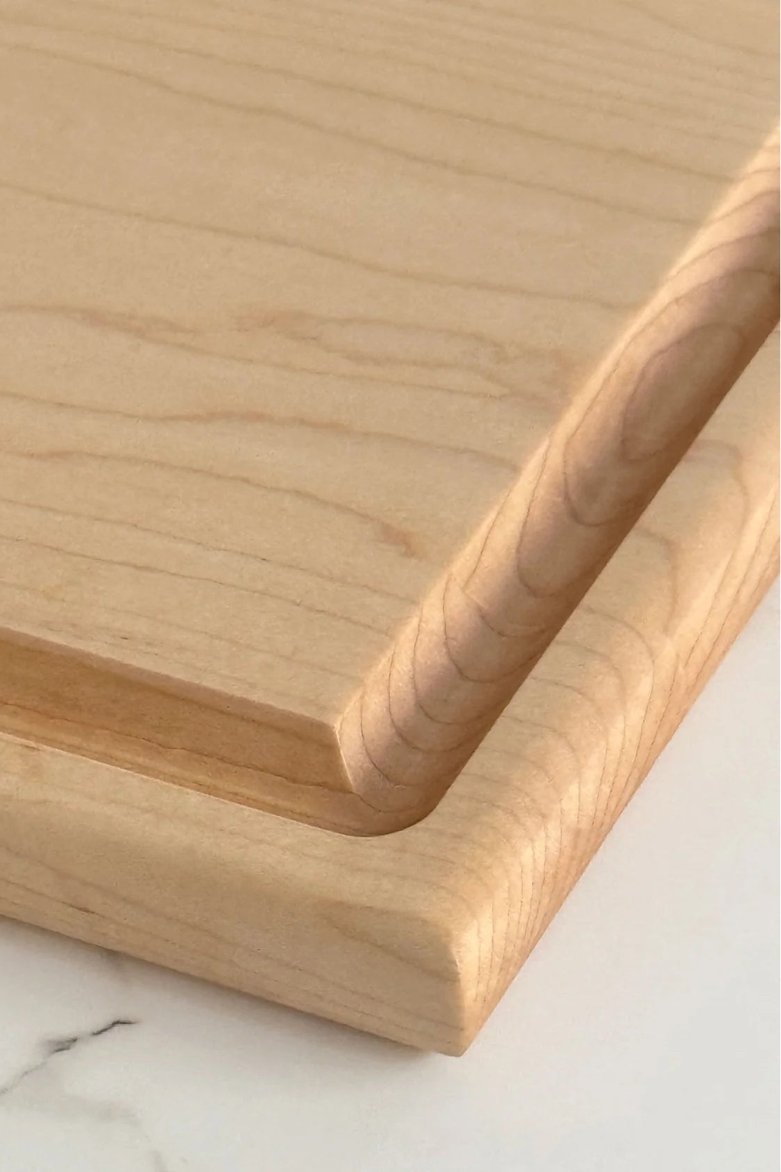 Close-up of a light natural wood cutting board with a recessed groove along one side, resting on a white marble surface.