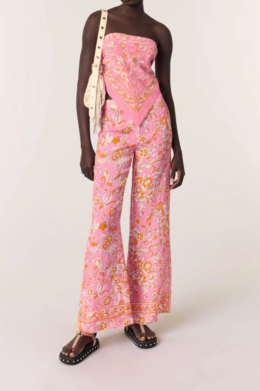A model stands against a plain light beige studio background wearing a pink strapless jumpsuit with a dense yellow and orange ornamental print, dark brown studded sandals, and a cream shoulder bag.