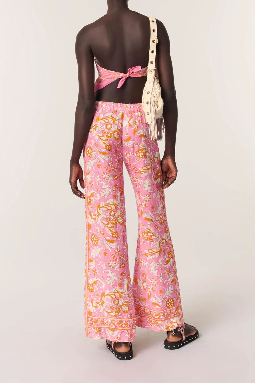 A model shown from the back wearing a pink floral wide-leg pants set with a matching pink bow-tie halter top, styled with a cream shoulder bag and black studded sandals against a plain light gray studio background.