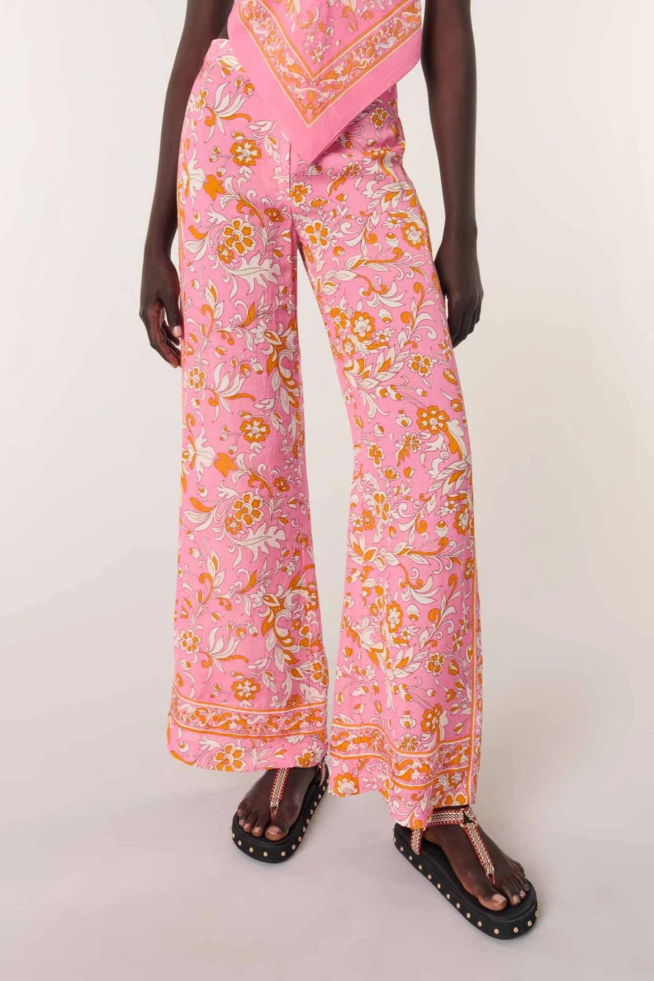 Pink wide-leg trousers with an orange and yellow floral paisley print, shown on a model wearing brown studded sandals against a light beige studio background.