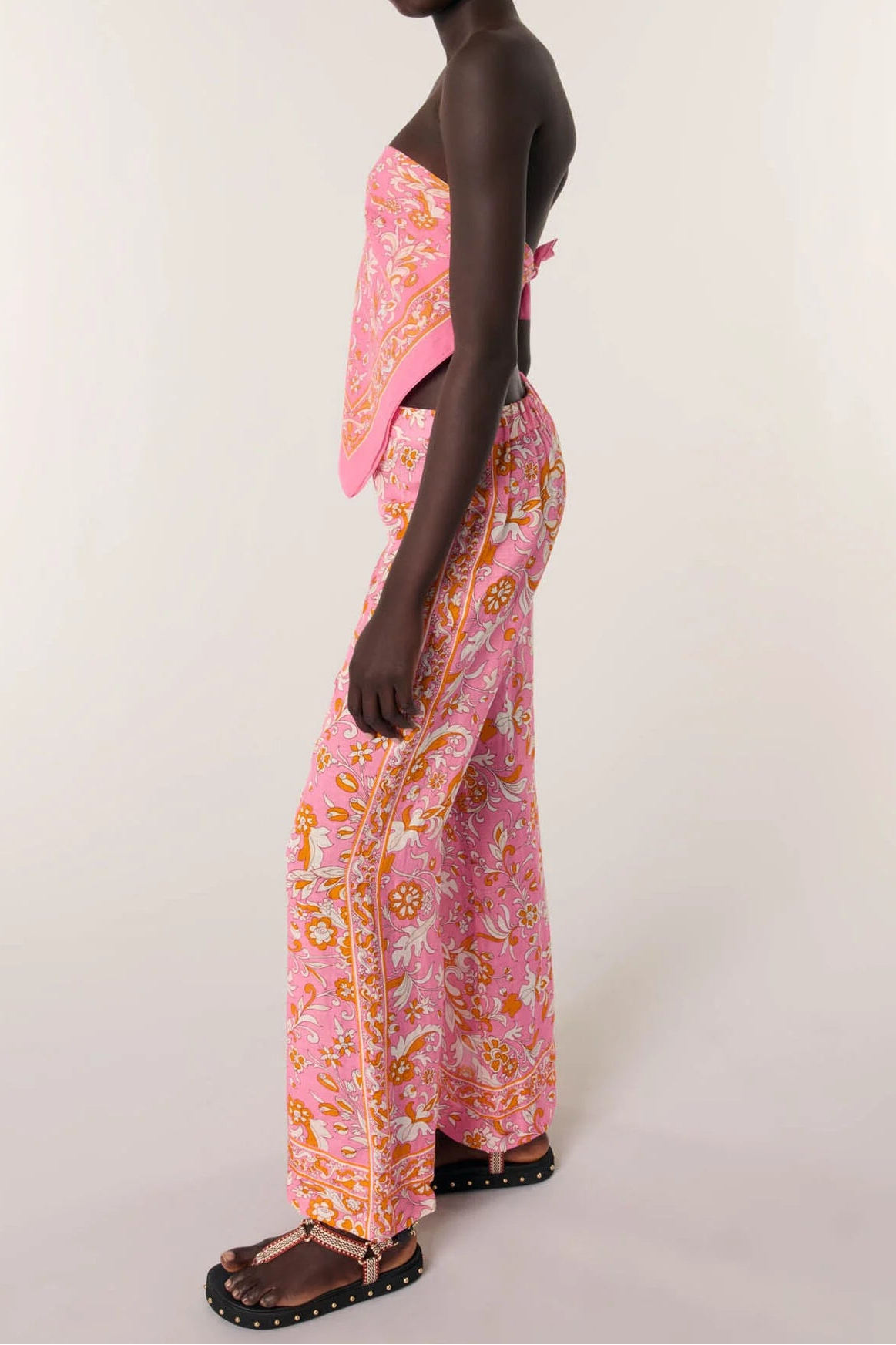 A model is shown from a side-back angle wearing a pink two-piece set with a colorful paisley pattern, consisting of a strapless top and matching wide-leg pants, against a plain light background.