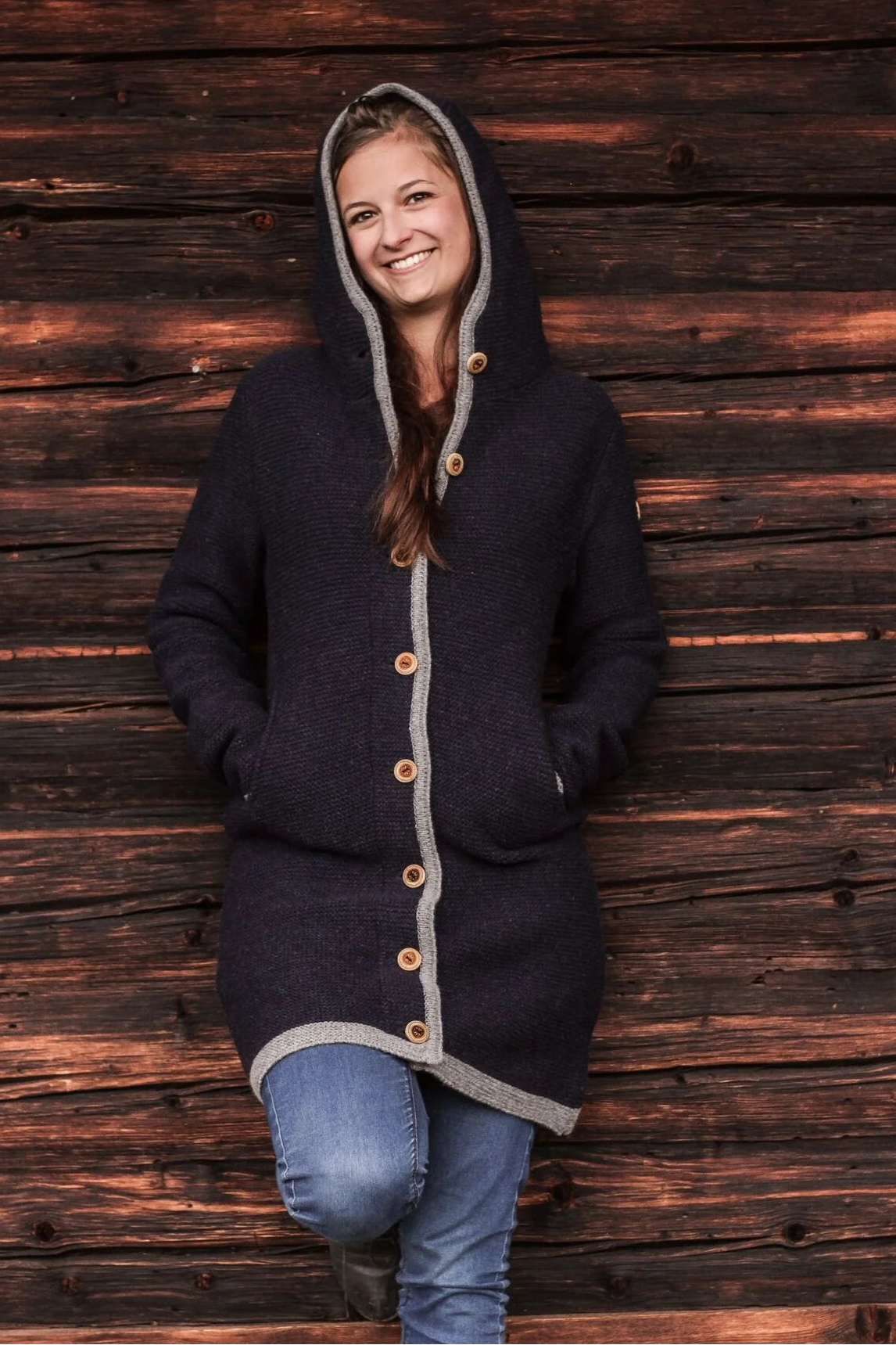 Woman wearing a dark blue hooded coat with wooden buttons against a wooden wall.