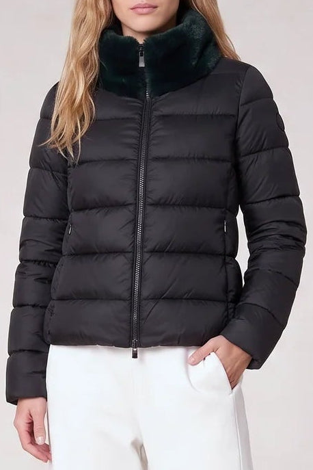 A woman stands against a plain light beige studio background wearing a black puffer jacket with a dark green faux-fur collar, white wide-leg trousers, and silver pointed shoes.