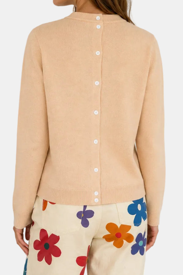 A woman modeling a light beige crewneck sweater, shown from both the front and back views against a white paneled wall. She is also wearing cream pants with colorful floral patterns.