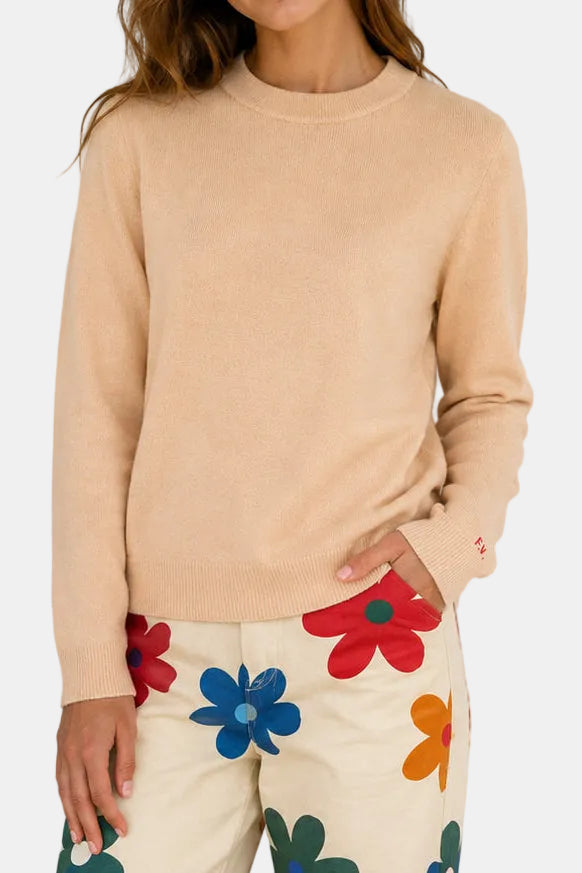 A woman modeling a light beige crewneck sweater, shown from both the front and back views against a white paneled wall. She is also wearing cream pants with colorful floral patterns.