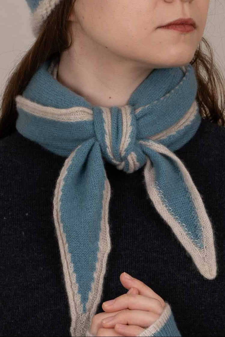 A close-up of a person wearing a knitted scarf tied in a bow around the neck. The scarf is light blue with cream-colored knit borders, and the person is wearing a dark navy sweater.