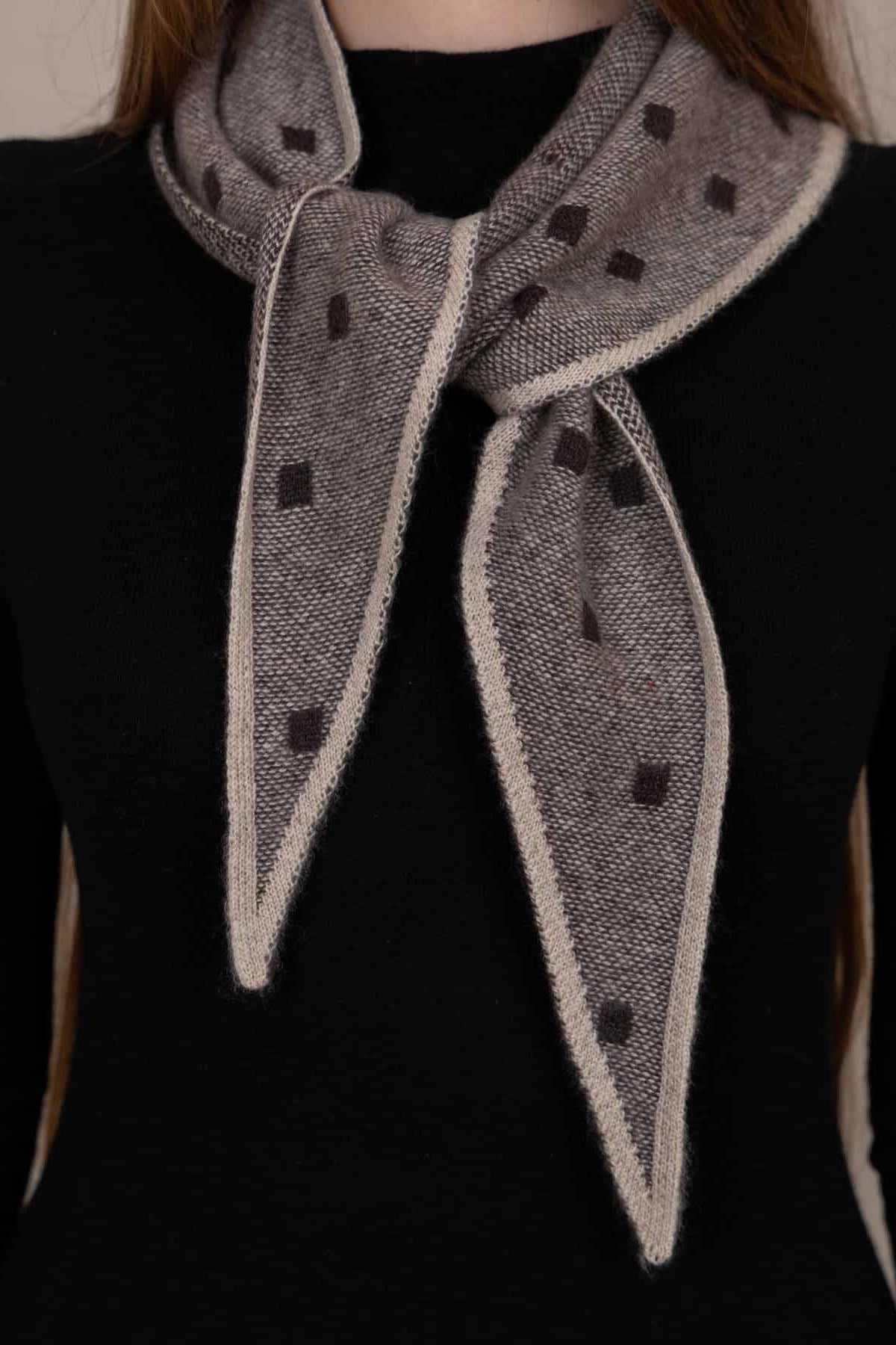 A close-up of a person wearing a gray knit scarf with dark brown polka dots and a cream border, tied around the neck over a black top.