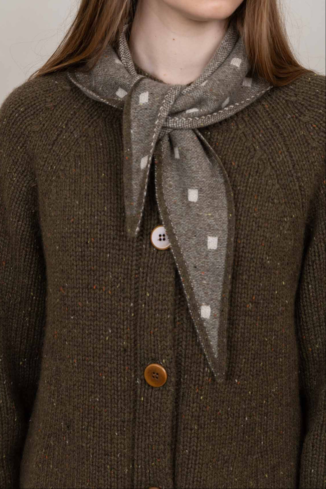 Close-up of a person wearing a dark brown textured knit cardigan with brown buttons and a gray patterned neck scarf tied at the collar.