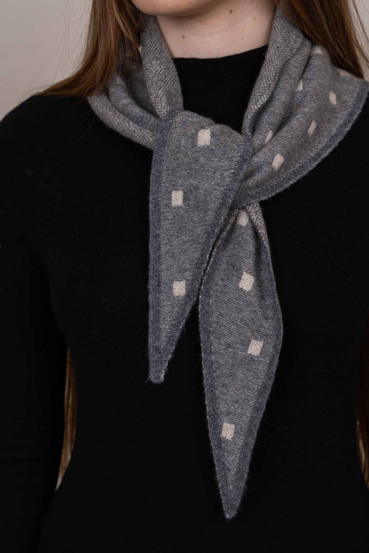 A close-up of a person wearing a gray knit scarf with small cream-colored square dots, tied around the neck over a black top.
