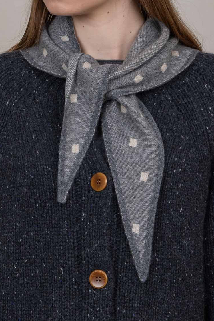 A woman wearing a dark navy knit beanie with a cream band, a gray neck scarf with cream polka dots, and a dark navy knit cardigan with brown buttons, photographed against a solid light gray background.