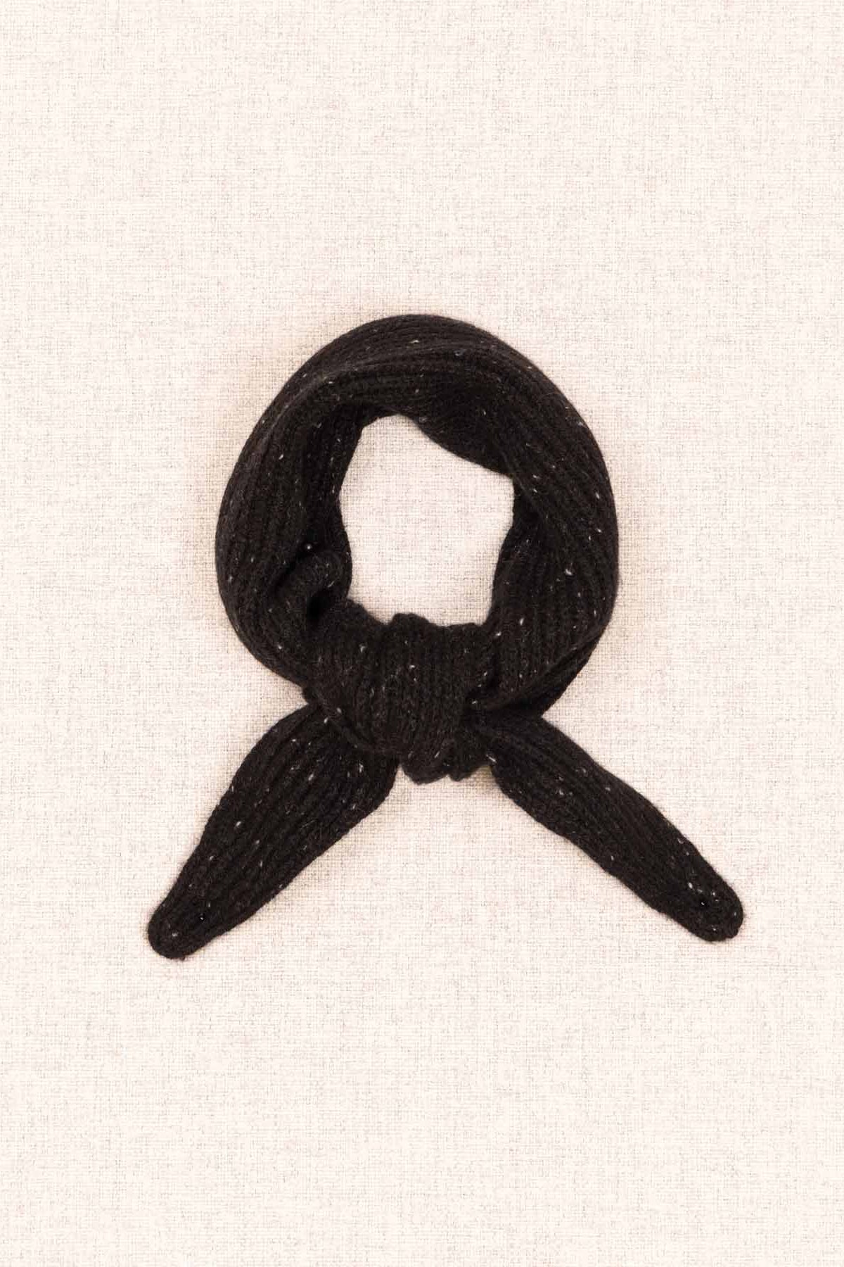 A small black knotted fabric accessory, possibly a miniature scarf, tie, or decorative knot, displayed on a textured off-white fabric background.
