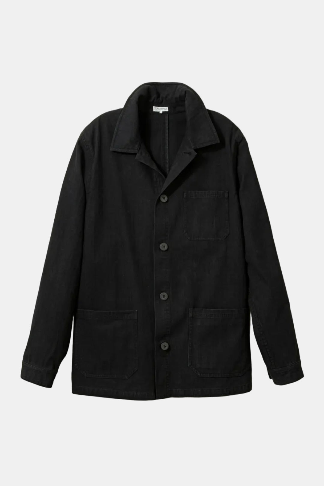 A black canvas utility jacket with a pointed collar, button-front closure, and four front pockets, displayed against a light beige wood-paneled background.