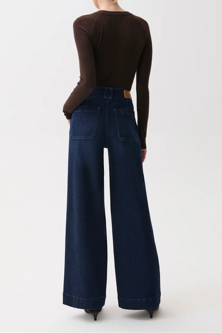 A woman shown from the back wearing a dark brown long-sleeve top and dark blue wide-leg denim jeans against a plain white studio background.