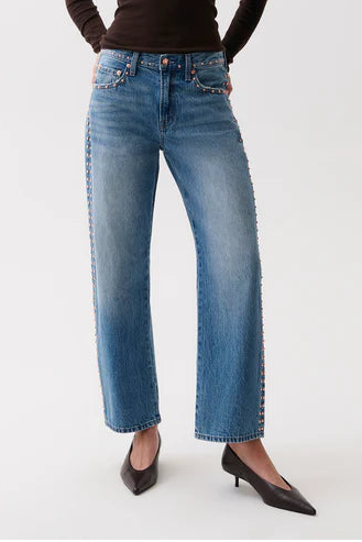 A woman modeling a dark brown long-sleeve knit top and wide-leg blue jeans with decorative silver studs down the sides, paired with black pointed-toe heels, against a plain white studio background.