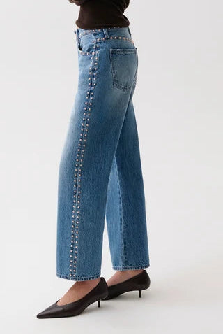 A woman modeling light-wash blue wide-leg jeans with silver chain detailing down the side, paired with a dark brown long-sleeve top and black pointed-toe heels, against a plain white studio background.