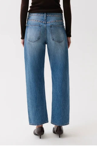 A woman viewed from behind wearing a dark brown long-sleeve top, blue wide-leg jeans, and black heels against a plain light gray studio background.
