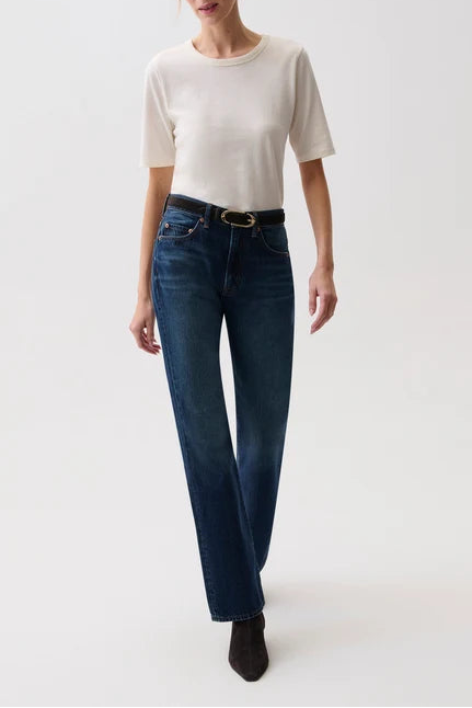A woman modeling a pair of dark blue straight-leg jeans with a black belt, paired with a cream short-sleeve t-shirt and black ankle boots, against a plain white studio background.