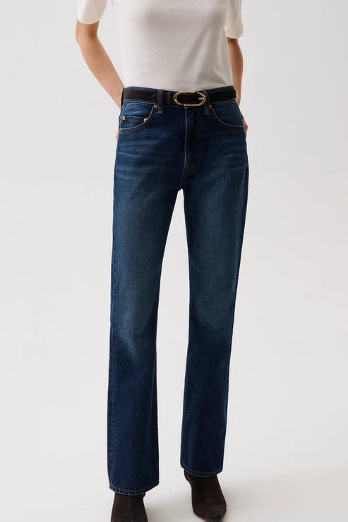 A model wearing dark blue straight-leg denim jeans with a black leather belt featuring a gold-tone oval buckle, styled with a white t-shirt and dark brown boots against a plain white studio background.