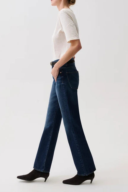 A woman in profile wearing dark blue wide-leg jeans, a white short-sleeve top, and black pointed-toe heeled boots, standing against a plain light gray studio background.