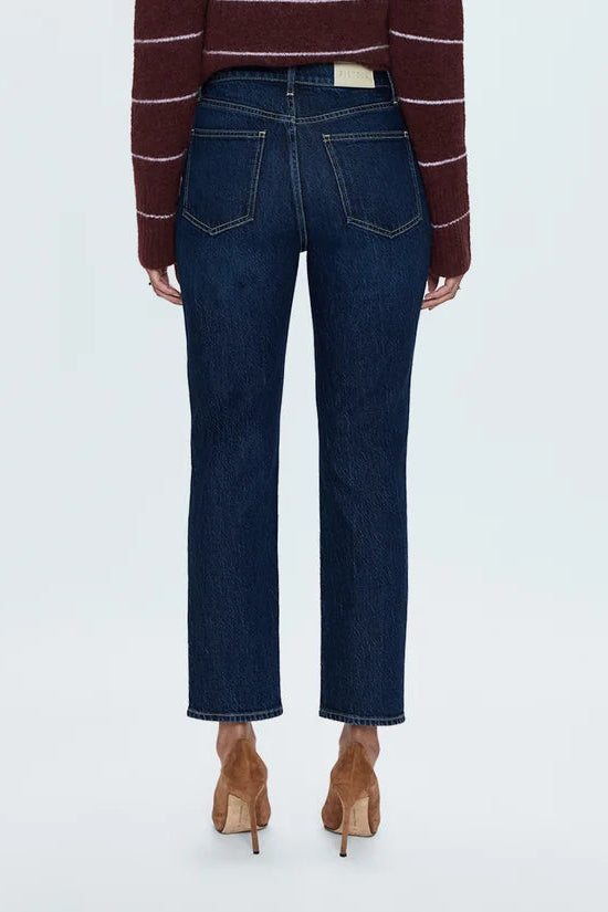A model viewed from the back wearing a dark burgundy cropped knit sweater with thin white horizontal stripes, paired with dark blue cropped jeans and brown high-heeled shoes, against a light gray studio background.