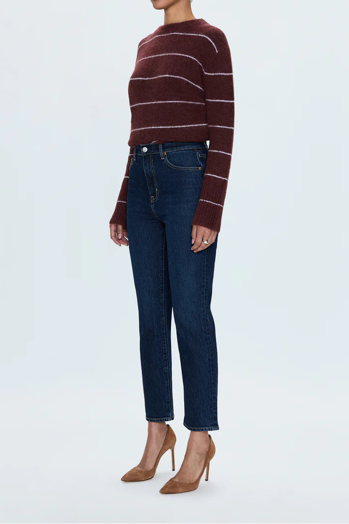 A woman modeling a dark brown and burgundy striped knit sweater paired with dark blue jeans and tan high heels, standing against a light gray studio background.