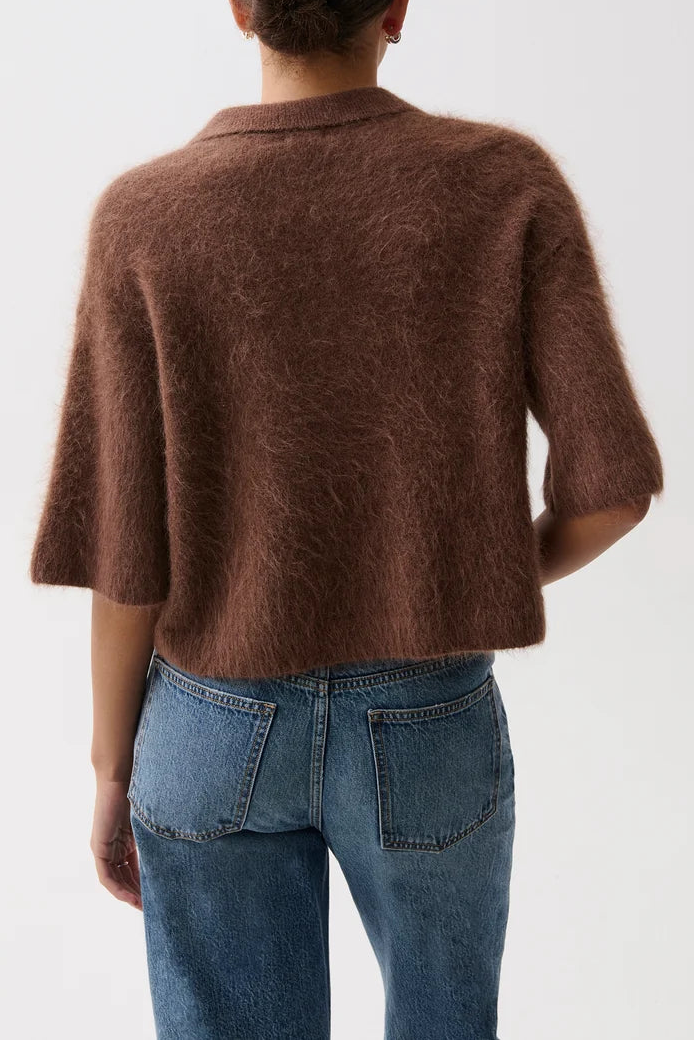 A model viewed from the back wearing a brown fuzzy short-sleeve cropped cardigan or sweater with blue jeans, set against a plain white studio background.