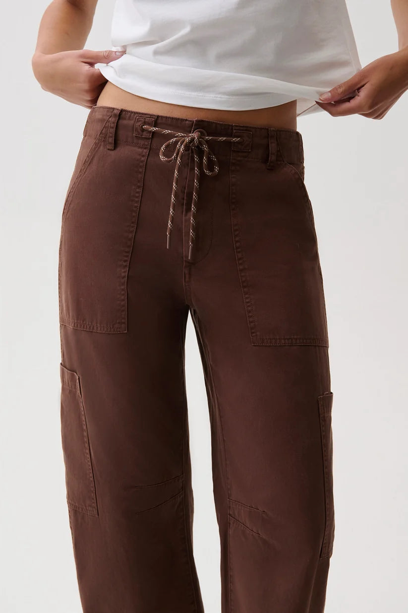 A close-up of a person wearing brown wide-leg cargo pants with a drawstring waistband, holding up a white t-shirt to show the fit.