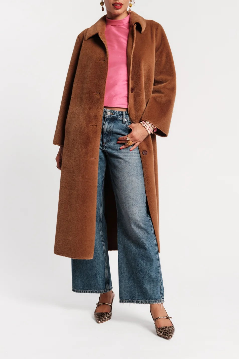 A model stands against a light gray studio background wearing a long brown suede-like coat over a pink turtleneck, blue wide-leg jeans, and leopard-print heels.